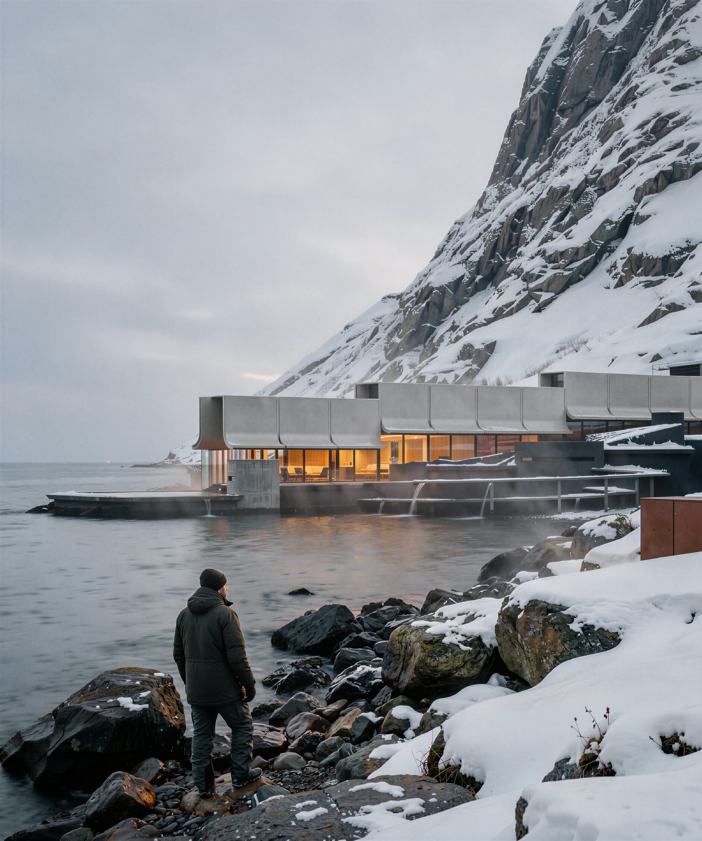 A figure in a winter coat on snow-covered rocks, looking towards the lit bathhouse beneath the mountain